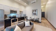 Located at 111 S Greenwood Ave in Tulsa, Oklahoma, this thoughtfully designed single-floor unit makes excellent use of its intimate footprint. Soaring ceilings throughout create a striking sense of verticality and openness that elevates the entire space.

The open-concept living and kitchen area serves as the heart of the unit. The kitchen is outfitted with stainless steel appliances, including a range, microwave, and refrigerator, paired with crisp white upper cabinetry and dark lower cabinetry for a modern two-tone contrast. A compact dining nook sits adjacent, ideal for casual meals. The living area accommodates comfortable seating and benefits from tall windows dressed in sheer curtains that invite abundant natural light. A wall-mounted mini-split HVAC system provides climate control with a clean, modern profile.

The bedroom is a calm, well-proportioned retreat with neutral walls and high ceilings that amplify the sense of space. Light wood-toned flooring runs throughout the main areas, adding warmth and continuity.

The bathroom features a clean, contemporary design with hexagonal white floor tile, a floating vanity with gold fixtures, a large mirror, and a full toilet. A sliding barn-style door adds an industrial-modern touch. The overall ambiance of the unit is modern and refined, with a neutral palette and curated finishes that balance style and functionality.
