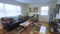 This single-floor residence offers a warm and inviting atmosphere with a thoughtfully arranged living space at its heart. The living room is anchored by rich hardwood floors that reflect natural light streaming in through multiple large windows fitted with white blinds, creating an airy and well-lit environment. The space comfortably accommodates a classic dark leather sofa and a coordinating gray upholstered armchair, both accented with decorative throw pillows. A wooden coffee table with metal legs sits atop a multicolored area rug, adding a layer of texture and warmth to the room. A side table nestled between the seating area provides additional surface space, while a decorative vase and dried botanicals add natural touches throughout. The walls are finished in a soft neutral tone, complemented by a vibrant abstract wall hanging that introduces a pop of color and artistic character. Wide baseboards and clean wall lines speak to the classic construction of the home. The overall ambiance is comfortable and functional, balancing everyday practicality with a welcoming aesthetic that makes the living area an ideal space for relaxing or entertaining.