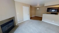 This single-story space features an open-concept layout that seamlessly connects the living and dining areas. The living area is finished with plush, light-toned carpeting, providing a comfortable and inviting atmosphere, while a fireplace with a sleek tile surround serves as a notable focal point along one wall. The flooring transitions into warm hardwood-style planks in the adjacent dining area, adding visual contrast and a sense of defined zones within the open floor plan.

A pendant light fixture hangs above the dining area, providing focused illumination and a subtle decorative touch. The walls are finished in a neutral beige tone throughout, contributing to a bright and cohesive aesthetic. White baseboards and trim offer clean contrast against the warm wall color.

A white six-panel door provides entry to the space and is framed neatly within the beige walls. Partially visible to one side is what appears to be a kitchen area with granite-style countertops, suggesting convenient adjacency to the main living spaces. The overall design is practical and straightforward, with a neutral palette that offers flexibility for a variety of interior styles.