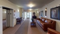 This single-floor lobby presents a well-appointed and hospitable environment designed to make a strong first impression. The entrance is anchored by a set of glass double doors that invite natural light into the corridor, offering views of the surrounding exterior. Patterned carpet in neutral tones runs the length of the space, adding texture and warmth underfoot.

The seating area is furnished with a rich cognac leather sofa and matching armchair, arranged around a solid wood coffee table adorned with a potted plant. This arrangement creates an inviting lounge atmosphere suitable for welcoming guests. A traditional dark wood bench with decorative spindle detailing is positioned near the entry doors, providing additional seating.

Wall-mounted sconces provide ambient lighting throughout, complemented by flush-mount ceiling fixtures. The walls are decorated with a curated selection of framed artwork and illuminated vertical display pieces, adding visual interest and a polished aesthetic. A sculptural wall installation featuring layered circular forms serves as a distinctive decorative accent near the entry.

A dark wood console table with decorative accessories further enhances the refined character of the space. Overall, the lobby conveys a comfortable, well-maintained atmosphere with thoughtful design details throughout.