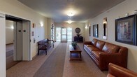 This single-floor lobby presents a well-appointed and hospitable environment designed to make a strong first impression. The entrance is anchored by a set of glass double doors that invite natural light into the corridor, offering views of the surrounding exterior. Patterned carpet in neutral tones runs the length of the space, adding texture and warmth underfoot.

The seating area is furnished with a rich cognac leather sofa and matching armchair, arranged around a solid wood coffee table adorned with a potted plant. This arrangement creates an inviting lounge atmosphere suitable for welcoming guests. A traditional dark wood bench with decorative spindle detailing is positioned near the entry doors, providing additional seating.

Wall-mounted sconces provide ambient lighting throughout, complemented by flush-mount ceiling fixtures. The walls are decorated with a curated selection of framed artwork and illuminated vertical display pieces, adding visual interest and a polished aesthetic. A sculptural wall installation featuring layered circular forms serves as a distinctive decorative accent near the entry.

A dark wood console table with decorative accessories further enhances the refined character of the space. Overall, the lobby conveys a comfortable, well-maintained atmosphere with thoughtful design details throughout.