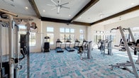 Located in Spring, TX, this fitness center is a single-floor facility designed to support a wide range of workout routines. The main workout area is expansive and architecturally distinguished by soaring vaulted ceilings with striking dark wood beam accents and large ceiling fans that keep air circulating throughout. A patterned teal and grey carpet tiles the floor, adding a modern, energetic aesthetic to the space. Floor-to-ceiling windows and large glass panels line the walls, inviting an abundance of natural light and offering views of the outdoor surroundings, including a waterfront amenity area visible from the cardio section. The main floor is well-stocked with a row of TRUE treadmills and ellipticals, HOIST cable and strength machines, rowing equipment, and additional cardio options. Wall-mounted televisions provide entertainment throughout. A dedicated studio room adjoins the main floor, featuring light hardwood flooring, full-length mirrors, a large flat-screen TV, spin bikes, resistance bands, medicine balls, yoga mats, and other functional training accessories neatly organized on built-in shelving. The entry area provides a welcoming transition into the facility, complete with reading material and wall-mounted displays.