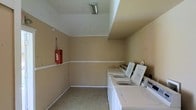This single-floor shared laundry facility is designed with practicality and functionality in mind. The room features a row of top-loading washing machines and dryers lined along one wall, providing ample capacity for multiple loads of laundry simultaneously. The space is finished with neutral beige walls accented by a white chair-rail molding that runs along the perimeter, lending a clean and tidy appearance. Overhead fluorescent lighting ensures the room is well-lit, making it easy to sort and manage laundry. The light-toned tile flooring is durable and easy to maintain, suited to the demands of a high-use utility space. A fire alarm pull station is visibly mounted on the wall, reflecting attention to safety standards. The layout is open and uncluttered, allowing for comfortable movement around the machines. Overall, this laundry room offers a straightforward, no-frills environment focused entirely on providing a convenient and accessible shared laundry solution.
