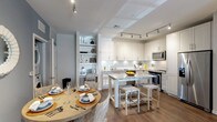 This single-floor residence is thoughtfully designed with an open-concept layout that seamlessly connects the kitchen, dining, and living areas. The kitchen is a standout feature, outfitted with white shaker cabinetry, granite countertops, a classic subway tile backsplash, and a full suite of stainless steel appliances. A granite-topped island with woven bar stools serves as both a prep space and casual dining area, while track lighting illuminates the room throughout. Adjacent to the kitchen, the dining area accommodates a round table with woven chairs and benefits from proximity to a stacked in-unit washer and dryer. The living room is bright and welcoming, featuring dark hardwood-style flooring, a ceiling fan, and a large window that allows natural light to fill the space. The bedroom is a calm retreat with neutral tones, plush carpet, natural light from a window, and ample room for full-sized furniture. The bathroom features a granite-topped vanity with grey cabinetry, light tile flooring, and a full soaking tub with tiled surround. Built-in open shelving in the living area provides additional storage and display space throughout.
