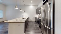 This single-floor residence showcases a thoughtfully designed open layout with contemporary finishes throughout. The kitchen serves as a standout feature, boasting an extended granite-topped island with an undermount sink and elegant veining that adds visual depth. Dark espresso cabinetry lines the walls, providing ample storage, while a herringbone tile backsplash adds a refined textural element. A suite of stainless steel appliances, including a French door refrigerator, range, and microwave, elevates the kitchen's functionality. Three sleek industrial-style pendant lights hang above the island, creating focused task lighting and an eye-catching design moment. Dark hardwood-style flooring flows throughout the main living and kitchen areas, offering a rich contrast to the bright white walls and tall ceilings that lend the space an open, airy feel. The bathroom mirrors the kitchen's design aesthetic with matching dark cabinetry, granite countertops, a wide vanity mirror, large-format tile flooring, and a separate glass-enclosed shower. A carpeted bedroom provides a warm, comfortable retreat, completing this well-balanced and cohesive living space.