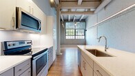 Located at 1587 E 31st St in Cleveland, OH, this one-bedroom, one-bathroom loft blends industrial character with contemporary design. The space retains its raw architectural roots through exposed white-painted brick walls, overhead ductwork, and beam-and-joist ceilings — all preserved to celebrate the building's industrial heritage. Warm hardwood flooring runs throughout, tying the open layout together cohesively.

The kitchen is a standout feature, offering stainless steel appliances including a refrigerator, electric range, and over-the-range microwave. Two-tone cabinetry — white upper cabinets paired with grey lowers — frames sleek quartz countertops, while a peninsula with an undermount sink provides additional prep and seating space. Track lighting illuminates the area effectively.

Large factory-style windows in the living area flood the space with natural light and offer views of surrounding greenery, enhancing the loft's airy, open feel. A ceiling fan complements the soaring ceiling height.

The bedroom features neutral grey walls, wood-look flooring, and a clean, minimalist aesthetic. The bathroom is finished with large-format grey floor tiles, a glass-enclosed walk-in shower with subway tile surround, and a quartz-topped vanity. A hallway with exposed brick connects the rooms, reinforcing the cohesive industrial-modern aesthetic throughout.