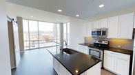 This single-floor space showcases a thoughtfully designed open-concept layout that seamlessly integrates the kitchen and living areas. The kitchen is a standout feature, anchored by a large central island with a deep single-basin sink and polished dark granite countertops that contrast elegantly against the white shaker-style cabinetry. Stainless steel appliances, including a range with oven and an over-the-range microwave, provide a sleek, modern aesthetic. Recessed lighting overhead ensures the kitchen remains well-lit and functional.

The space transitions effortlessly toward an expansive wall of floor-to-ceiling windows that frame commanding views of the surrounding urban skyline. Natural light floods the interior, highlighting the clean white walls and light-toned flooring. The open floor plan creates an airy, spacious atmosphere well-suited for both daily living and entertaining. The minimalist design philosophy is consistent throughout, with a neutral color palette that allows the dramatic cityscape views and premium finishes to take center stage, giving the space a polished, contemporary character.