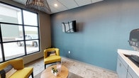 This single-floor space presents a thoughtfully designed lounge or waiting area with a clean, contemporary aesthetic. The room is defined by a striking blue-gray accent wall, which serves as a bold focal point and anchors the seating arrangement. A wall-mounted flat-screen television is positioned prominently on this accent wall, adding a functional element to the space.

The seating area features vibrant yellow accent chairs that provide a lively contrast against the cooler tones of the walls, creating an inviting and visually dynamic environment. A round wooden coffee table sits at the center, styled with small potted succulents and decorative accents that add a touch of warmth and life to the space.

Natural light enters through a large window along one wall, offering views to the exterior and contributing to a bright, open atmosphere. The drop tile ceiling is complemented by a distinctive sculptural pendant light fixture with a starburst design, lending an artistic touch to the overhead plane.

The flooring consists of neutral-toned carpet, and the overall color palette—combining soft grays, muted blues, and warm yellow accents—creates a balanced, professional yet comfortable ambiance suitable for receiving guests or providing a relaxed waiting experience.
