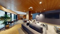 This thoughtfully designed amenity lounge seamlessly blends entertainment and relaxation across a single open-plan floor. The space is defined by a striking warm wood-paneled ceiling that spans much of the interior, complemented by recessed lighting and wood-clad columns that subtly divide the room into distinct functional zones without sacrificing its open, cohesive feel.

The media lounge anchors one end of the space, featuring generously sized gray sectional sofas arranged around a wall-mounted flat-screen television. A deep blue textured accent wall adds depth and contrast, while a marble-topped coffee table grounds the seating arrangement.

Adjacent to the lounge, a games area houses a full-sized pool table beneath a large pendant light, alongside a shuffleboard table, both set atop a patterned tile floor with a star motif. A wet bar with dark cabinetry and stone countertops provides convenient service nearby. A cozy dining nook with warm-toned chairs and framed artwork offers a more intimate setting.

Floor-to-ceiling windows along one wall flood the space with natural light and provide views of an adjoining outdoor area. The overall ambiance is sophisticated yet inviting, balancing modern design elements with comfortable, social-oriented furnishings.