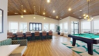 This well-appointed single-floor community clubhouse is designed for socializing and recreation, combining modern aesthetics with comfortable, functional spaces throughout. The main gathering area is anchored by soaring vaulted ceilings clad in warm wood planking, with pendant globe lighting that adds a contemporary touch. Light hardwood flooring runs through the open-plan layout, which flows seamlessly between a games area, dining spaces, and a fully equipped kitchen.

The games room features a full-size billiards table set atop a bold teal-and-cream geometric area rug, surrounded by large windows that flood the space with natural light and offer views of an adjoining outdoor balcony. The kitchen is generously sized, with an extended island featuring green cabinetry and white countertops, bar-height seating, stainless steel appliances, and a wine cooler — ideal for catering events or casual gatherings.

A dedicated lounge area offers a plush sectional sofa on richly patterned carpet, alongside a wall-mounted television, creating a cozy media nook. A stylish wet bar with a mirrored mosaic tile backsplash and under-counter wine refrigerator adds an elegant entertaining option. The entryway features glass-panel doors, high ceilings, and additional café-style seating. Throughout, a cohesive teal, white, and wood color palette ties the space together with a fresh, inviting ambiance.
