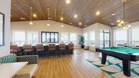 This well-appointed single-floor community clubhouse is designed for socializing and recreation, combining modern aesthetics with comfortable, functional spaces throughout. The main gathering area is anchored by soaring vaulted ceilings clad in warm wood planking, with pendant globe lighting that adds a contemporary touch. Light hardwood flooring runs through the open-plan layout, which flows seamlessly between a games area, dining spaces, and a fully equipped kitchen.

The games room features a full-size billiards table set atop a bold teal-and-cream geometric area rug, surrounded by large windows that flood the space with natural light and offer views of an adjoining outdoor balcony. The kitchen is generously sized, with an extended island featuring green cabinetry and white countertops, bar-height seating, stainless steel appliances, and a wine cooler — ideal for catering events or casual gatherings.

A dedicated lounge area offers a plush sectional sofa on richly patterned carpet, alongside a wall-mounted television, creating a cozy media nook. A stylish wet bar with a mirrored mosaic tile backsplash and under-counter wine refrigerator adds an elegant entertaining option. The entryway features glass-panel doors, high ceilings, and additional café-style seating. Throughout, a cohesive teal, white, and wood color palette ties the space together with a fresh, inviting ambiance.