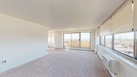 Located at 2400 McClellan Ave in Pennsauken, NJ, this elevated unit offers a bright and open layout with sweeping panoramic views of the surrounding landscape, including distant city skyline glimpses. The spacious living area is defined by wall-to-wall carpeting and an impressive bank of large windows fitted with roller shades, allowing abundant natural light to fill the room. A sliding glass door opens directly onto a private balcony, further extending the sense of openness and providing an ideal vantage point for the surrounding views. Baseboard heating and a wall-mounted air conditioning unit ensure year-round comfort throughout the living space. The kitchen is thoughtfully equipped with warm wood cabinetry, granite-style countertops, a gas range, dishwasher, and refrigerator, all set on easy-care tile flooring. The floor plan, as seen in the dollhouse view, includes multiple private rooms, offering flexible use as bedrooms or additional living space, each finished with carpet and large windows. Two bathrooms are present, both finished with classic mosaic tile flooring and clean white fixtures, one featuring a shower-tub combination. The overall aesthetic is clean, neutral, and move-in ready, with a light color palette throughout.