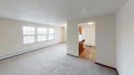 This single-floor unit offers a straightforward and functional layout, as illustrated by the 3D floor plan. The living area is generously sized, featuring neutral-toned carpet, cream-colored walls, and a row of windows fitted with horizontal blinds that allow ample natural light to fill the space. A baseboard heating system runs along the perimeter, and the textured ceiling adds subtle dimension. The living area connects openly to the kitchen, which is separated by a half-wall partition. The kitchen features warm oak cabinetry—both upper and lower—paired with neutral laminate countertops, wood-look flooring, and white appliances including a refrigerator and electric range with overhead ventilation. The bedrooms are carpeted and well-proportioned, each with a window, baseboard heat, and a wall-mounted AC unit in one room for individual climate control. The bathroom includes wood-look vinyl flooring, an oak vanity with a single sink, a wall-mounted mirror with overhead lighting, a storage cabinet, and a tub/shower combination with a curtain rod. Overall, the unit presents a clean, practical living environment with consistent warm-toned wood accents throughout.