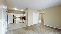 This single-floor residence offers a practical and well-organized open-concept layout that seamlessly connects the kitchen and living areas. The kitchen is fully equipped with white cabinetry, a gas range, built-in microwave, dishwasher, and dark countertops, all arranged in an efficient L-shaped configuration. Ample upper and lower cabinet storage provides generous space for kitchen essentials. The flooring transitions from easy-care hard flooring in the kitchen to plush wall-to-wall carpeting in the living area, creating a comfortable and cohesive atmosphere throughout. Warm beige walls contribute to a neutral, inviting ambiance that complements a variety of décor styles. A large double-door closet is conveniently located near the living area, offering substantial storage capacity. A hallway leads from the main living space to additional rooms, suggesting a well-considered floor plan designed for everyday functionality. Ceiling-mounted light fixtures provide consistent illumination throughout, and multiple HVAC vents ensure climate comfort year-round. Overall, the space delivers a clean, move-in-ready environment with a straightforward layout suited for comfortable everyday living.