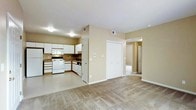 This single-floor residence offers a practical and well-organized open-concept layout that seamlessly connects the kitchen and living areas. The kitchen is fully equipped with white cabinetry, a gas range, built-in microwave, dishwasher, and dark countertops, all arranged in an efficient L-shaped configuration. Ample upper and lower cabinet storage provides generous space for kitchen essentials. The flooring transitions from easy-care hard flooring in the kitchen to plush wall-to-wall carpeting in the living area, creating a comfortable and cohesive atmosphere throughout. Warm beige walls contribute to a neutral, inviting ambiance that complements a variety of décor styles. A large double-door closet is conveniently located near the living area, offering substantial storage capacity. A hallway leads from the main living space to additional rooms, suggesting a well-considered floor plan designed for everyday functionality. Ceiling-mounted light fixtures provide consistent illumination throughout, and multiple HVAC vents ensure climate comfort year-round. Overall, the space delivers a clean, move-in-ready environment with a straightforward layout suited for comfortable everyday living.