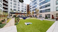Located at 616 N Glebe Road in Arlington, Virginia, this outdoor courtyard amenity space is set within the interior of a contemporary multi-story residential building. The courtyard is thoughtfully designed to offer a variety of recreational and social environments, all centered around a broad, well-maintained artificial turf lawn that serves as the focal point of the space.

Surrounding the lawn, distinct zones offer different experiences. A sleek pergola structure with a flat canopy roof anchors one side of the courtyard, sheltering an outdoor lounge and dining area furnished with chairs and tables, ideal for gatherings. Overhead string lighting extends across portions of the space, adding ambiance during evening hours.

Adjacent to the covered seating area, an outdoor grilling station equipped with built-in appliances provides a functional cooking area. Casual blue pouf seating is scattered across the lawn area, and cornhole boards are set up for recreational use. A wood-deck walkway runs along one side, offering a transitional surface between zones.

Landscaping throughout features manicured shrub borders, ornamental trees with seasonal color, natural stone retaining walls with granite-topped ledges, and layered plantings that add texture and visual interest. The surrounding building's exterior combines white horizontal siding, brick, and warm wood-toned cladding, framing the courtyard on all sides and creating an enclosed, community-oriented atmosphere.