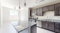 This contemporary single-floor unit offers a clean, modern aesthetic defined by a neutral color palette, light hardwood-style flooring, and an abundance of natural light. The kitchen is a standout feature, showcasing rich dark espresso-toned shaker cabinetry that extends to the ceiling, providing ample storage. A large central island with a striking grey-veined granite countertop serves as both a workspace and casual gathering area, illuminated by stylish cylindrical pendant lights overhead. A full suite of stainless steel appliances, including a refrigerator, range, microwave, and dishwasher, completes the kitchen with a polished, functional finish. The open layout connects the kitchen seamlessly to the living and dining areas, where a ceiling fan and large windows invite generous sunlight and views of the surrounding neighborhood. The living area features high ceilings that enhance the sense of openness. The bedrooms are bright and airy, each benefiting from large windows that frame outdoor views of the community. Light-toned walls and continuous hardwood-style flooring unify the space, creating a cohesive and inviting atmosphere throughout. A closet with sliding panel doors adds practical storage solutions to the unit.
