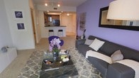 This single-floor residence showcases a thoughtfully designed open-concept layout that connects the living and kitchen areas into one cohesive, functional space. The living room is anchored by a charcoal gray sofa dressed with plush accent pillows and a cozy throw, positioned across from a glass-topped coffee table resting on a patterned area rug. A vibrant purple accent wall adds a bold, contemporary touch to the space, complemented by a large decorative mirror and tasteful wall art that enhance the room's visual depth. A purple accent chair adds a playful pop of color, tying the design palette together cohesively.

The kitchen, visible from the living area, features dark cabinetry with a raised breakfast bar outfitted with modern bar stools, creating a casual dining option. Track lighting illuminates the kitchen, while recessed ceiling lights provide ambient brightness throughout the open living space. Neutral-toned carpeting runs through the main living area, contributing warmth and texture underfoot. The overall ambiance of the space is modern and vibrant, balancing bold color choices with clean lines and contemporary furnishings to create an inviting, stylish environment.