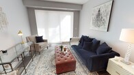 This thoughtfully designed single-floor residence offers a seamless blend of modern aesthetics and comfortable living. The open-concept layout connects the living, dining, and kitchen areas, creating a cohesive and functional space throughout.

The living area is anchored by rich, dark hardwood flooring and soft grey-toned walls, providing a sophisticated backdrop for the carefully curated furnishings. A large sliding panel window floods the space with natural light, highlighting key features such as a plush navy velvet sofa, a tufted leather ottoman, and a patterned area rug. A glass-and-metal console table and framed wall art add refined decorative touches.

The kitchen is a standout feature, boasting an expansive white quartz peninsula with an undermount sink, crisp white cabinetry extending to the ceiling, and a full suite of stainless steel appliances including a gas range, built-in microwave, and French door refrigerator. Pendant lighting overhead adds a contemporary finishing detail.

The bedroom is a serene retreat, featuring the same warm hardwood flooring, a large window with roller shade for natural light, and ample storage provided by a substantial grey dresser. The bathroom is finished with classic white subway tile surrounding the tub-shower combination, complemented by clean grey walls and chrome fixtures throughout.