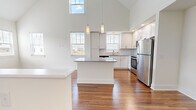 This two-story home in Lebanon, Indiana is designed with a bright, open layout spread across two well-proportioned floors. The main level features a dramatic vaulted ceiling that rises above the combined kitchen and living area, creating an airy, expansive atmosphere enhanced by abundant natural light from multiple windows. The kitchen is well-appointed with white shaker cabinetry extending to the ceiling, granite countertops, a central island, stainless steel appliances including a range, microwave, dishwasher, and refrigerator, and warm hardwood flooring throughout. Pendant lighting above the island adds a refined touch. The living area is adjacent to the staircase, which leads to the upper level. Also on the main floor, the full bathroom features granite countertops, white cabinetry, and hardwood flooring, with a dedicated walk-in closet nearby. The bedroom is carpeted and includes a ceiling fan and a window with neighborhood views. The upper level offers a sizable additional carpeted room with a sloped ceiling, providing flexible bonus space suitable for a home office or den.
