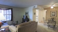 This single-floor residence offers a straightforward and functional layout designed for comfortable everyday living. The living room serves as the central gathering space, featuring wall-to-wall carpeting that adds warmth underfoot. The room provides ample space for seating and entertainment furniture, and is finished with a textured ceiling and neutral-toned walls that create a calm, understated ambiance. A HVAC vent is integrated into the ceiling, ensuring year-round climate control. An open passageway connects the living area to the kitchen, which is visible in the background and equipped with essential appliances including a range. The kitchen features light cabinetry and a clean, efficient workspace. The transition between the two spaces is defined by a structural soffit overhead, adding architectural character to the open-plan feel. Natural and artificial lighting work together to keep the interior bright. The entry door is accessible directly from the living room, adding convenience to the overall flow of the unit. The consistent use of neutral tones and carpeted flooring throughout ties the spaces together cohesively, making this a welcoming and practical residential space.