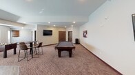 This single-floor recreation and community room is thoughtfully designed to offer a variety of leisure and social activities within one open, well-appointed space. The room features neutral white and beige walls complemented by patterned carpet throughout the main areas, transitioning to wood-look flooring near the kitchen zone, creating a clear and functional distinction between spaces.

The game area serves as a central focal point, anchored by a full-size pool table with dark wood legs and felt surface. A foosball table nearby adds additional entertainment, while wall-mounted flat-screen televisions are positioned for easy viewing from multiple areas of the room. Colorful billiards-themed artwork adorns the walls, reinforcing the playful character of the space. High-top tables with bar-style chairs are arranged throughout, offering casual seating for guests.

The kitchen area is fully equipped with stainless steel appliances, including a refrigerator, range, and microwave, set beneath rich dark-stained cabinetry with granite countertops. A classic red popcorn machine adds a charming, festive touch near the kitchen entry. Recessed lighting throughout ensures the entire space is well-lit, and access doors lead to additional rooms and exits, providing convenient flow and accessibility.