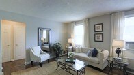 This single-floor space presents a well-appointed living room designed with a clean, neutral aesthetic. The room is finished with light grey walls and a textured ceiling, contributing to a calm and cohesive atmosphere. Natural light enters through a large window dressed with elegant white curtains, brightening the space and creating an airy feel.

The living area is furnished with a cream-colored sofa accented by navy and white throw pillows, paired with a patterned accent chair that adds subtle visual interest. A glass-topped coffee table with a dark metal frame serves as a central focal point, resting atop a natural woven area rug that warms the hardwood flooring beneath.

A large floor mirror with a metallic frame is positioned along one wall, effectively amplifying the sense of space and reflecting light throughout the room. A potted indoor plant introduces a touch of greenery, while framed artwork on the opposite wall adds a finishing decorative detail. A table lamp near the window provides warm supplemental lighting. Overall, the space balances comfort and style with a thoughtfully curated, understated elegance.