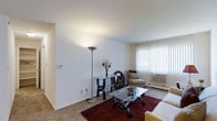 This single-floor residential unit offers a clean, functional layout designed for comfortable everyday living. The living room is finished with wall-to-wall carpeting and features smooth white walls and a flat ceiling that lend the space a bright, open feel. A large window fitted with vertical blinds allows natural light to fill the room while providing privacy, and a wall-mounted air conditioning unit ensures year-round comfort. The living area provides ample room for a full seating arrangement and accent furnishings, creating a welcoming and versatile space.

Beyond the interior, residents have access to a generously sized outdoor swimming pool, enclosed by a wrought iron fence and surrounded by a wide pool deck lined with lounge chairs — ideal for relaxing during warmer months. The pool area is framed by mature willow and shade trees, with well-maintained green lawns stretching toward the neighboring multi-story residential buildings. This combination of a comfortable interior and attractive outdoor amenities makes the space well-suited for those seeking a balanced residential environment with both private living quarters and shared community features.
