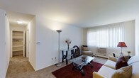 This single-floor residential unit offers a clean, functional layout designed for comfortable everyday living. The living room is finished with wall-to-wall carpeting and features smooth white walls and a flat ceiling that lend the space a bright, open feel. A large window fitted with vertical blinds allows natural light to fill the room while providing privacy, and a wall-mounted air conditioning unit ensures year-round comfort. The living area provides ample room for a full seating arrangement and accent furnishings, creating a welcoming and versatile space.

Beyond the interior, residents have access to a generously sized outdoor swimming pool, enclosed by a wrought iron fence and surrounded by a wide pool deck lined with lounge chairs — ideal for relaxing during warmer months. The pool area is framed by mature willow and shade trees, with well-maintained green lawns stretching toward the neighboring multi-story residential buildings. This combination of a comfortable interior and attractive outdoor amenities makes the space well-suited for those seeking a balanced residential environment with both private living quarters and shared community features.