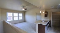 This 2-floor space showcases an open-concept layout that seamlessly connects the living area and kitchen. The living room features vaulted ceilings that enhance the sense of spaciousness, plush wall-to-wall carpeting, and a ceiling fan with integrated lighting. Natural light filters in through dual windows, brightening the neutral-toned walls and creating an airy atmosphere.

The kitchen is positioned adjacent to the living area and is distinguished by a raised breakfast bar counter with light stone-look countertops, a stainless steel sink, and rich dark-toned cabinetry. Two sleek pendant lights hang above the counter, providing focused task lighting and adding a modern design element to the space.

A doorway visible from the living area suggests additional rooms beyond, contributing to the functional flow of the floor plan. The overall design palette—neutral walls, dark cabinetry, and light countertops—creates a clean, contemporary aesthetic. The combination of vaulted ceilings, open sightlines, and thoughtful lighting makes this a welcoming and well-appointed multi-floor space.