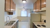 This single-floor space centers around a practical, galley-style kitchen equipped with warm wood cabinetry that lines both the upper and lower walls, providing generous storage throughout. The kitchen features a stainless steel sink with adjacent counter space, a dishwasher, a freestanding refrigerator, and an electric range, offering all the essentials for everyday cooking. Light-toned countertops complement the cabinetry, while the light-colored tile flooring runs seamlessly throughout the space, contributing to a bright and cohesive feel. A textured ceiling with a flush-mount light fixture illuminates the kitchen area. The space opens into an adjoining room, likely a dining or living area, which benefits from natural light streaming through a window fitted with blinds. A ceiling fan positioned in this area enhances air circulation and adds a functional decorative element. A baseboard heater along the wall ensures year-round comfort, while a wall-mounted air conditioning unit provides cooling. Wood-toned baseboards and trim detail the transition between rooms, adding a subtle finishing touch throughout the space.