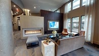 This single-story space showcases a refined, contemporary design anchored by a striking linear gas fireplace set within a textured stone surround. A rich, dark wood accent wall frames a wall-mounted flat-screen television, serving as a bold focal point for the lounge area. The soaring double-height ceilings amplify the sense of openness, while large floor-to-ceiling windows along the perimeter flood the interior with abundant natural light and offer views of the surrounding greenery outside.

The seating area is thoughtfully arranged with a generously sized sectional sofa in warm earth tones, complemented by a cognac leather accent chair and a sleek, upholstered ottoman. A glass-top coffee table and a stone-finished side cube add layered texture and visual interest throughout the space. Recessed lighting installed in the ceiling provides subtle ambient illumination, enhancing the warm, welcoming atmosphere.

The neutral color palette—incorporating tones of taupe, brown, and grey—alongside the mix of natural materials such as stone, wood, and leather, creates a cohesive and sophisticated ambiance suited for relaxation and social gatherings.