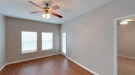 This single-floor space showcases a clean, functional design with warm hardwood flooring that runs throughout the main living area, adding a sense of warmth and continuity. The neutral gray walls provide a versatile backdrop, well-suited for a variety of interior styles and furnishing choices. A ceiling fan with an integrated light fixture serves as a central focal point overhead, offering both practical air circulation and ambient lighting. Multiple windows dressed with white horizontal blinds allow generous natural light to filter into the room, contributing to an open and airy feel. A glass-paneled door provides additional light and likely leads to an outdoor area. White baseboards frame the perimeter of the room, offering a crisp contrast to the flooring and walls. The space is open and unfurnished, allowing a clear view of its layout and proportions, which suggest comfortable, flexible use. Overall, the room presents a tidy, move-in-ready environment with straightforward, practical finishes throughout.