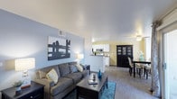 This single-floor residence offers an open-concept layout that seamlessly connects the living, dining, and kitchen areas into one cohesive and functional space. The living room features light blue accent walls, smooth white ceilings, and wood-style flooring that extends throughout, creating a clean and contemporary feel. A comfortable seating area anchors the room, complemented by a bold abstract wall piece that adds a artistic focal point. Natural and ambient lighting work together to keep the space bright and inviting.

The dining area transitions naturally from the living space and is highlighted by a ceiling fan overhead, a dark wood china cabinet, and a defined dining set that adds a touch of classic contrast against the lighter surroundings. The kitchen, visible from the living room via a pass-through counter, features white cabinetry, stainless steel appliances including a microwave, and a cheerful yellow accent wall that lends warmth and energy to the cooking space. The open sightlines between all three areas make the floor plan feel spacious and connected, ideal for both everyday living and entertaining. Overall, the space balances modern simplicity with comfortable, functional design.