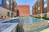 Located at 520 Martin Luther King Jr Blvd in downtown Macon, Georgia, this amenity space offers a well-rounded combination of outdoor recreation and indoor fitness facilities. The centerpiece of the outdoor area is a long, resort-style swimming pool flanked by modern multi-story buildings with a striking mix of wood, brick, and metal facades. Orange lounge chairs line the poolside deck, and an eye-catching decorative water feature wall serves as a bold architectural backdrop. Lush greenery and paved walkways complete the inviting courtyard setting.

The fitness center is thoughtfully divided into distinct functional zones. A cardio area is outfitted with True-brand treadmills, ellipticals, and stationary bikes, positioned in front of large picture windows that overlook the streetscape, bringing in abundant natural light and views of the surrounding neighborhood. A dedicated strength training section houses Freemotion cable machines and a multi-functional weight rack stocked with kettlebells, medicine balls, a squat rack, and benches. Rubber flooring throughout provides durability and safety. The facility is finished with neutral-toned walls, ceiling fans for air circulation, and a clean, functional aesthetic suited for a comprehensive daily workout routine.
