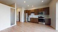 This single-floor unit presents a contemporary open-concept design where the kitchen and living area flow seamlessly together. The kitchen is a standout feature, outfitted with rich walnut-toned flat-front cabinetry extending from floor to ceiling, providing generous storage throughout. A white herringbone-patterned tile backsplash adds visual texture, while light quartz countertops offer a clean contrast. A full suite of stainless steel appliances — including a refrigerator, dishwasher, microwave, and range — ensures modern functionality. A built-in wine rack adds a refined detail above the sink area.

The living space is bright and open, with light hardwood-style flooring running throughout. A large sliding glass door fitted with vertical blinds floods the area with natural light and provides outdoor views. The overall palette of warm whites and neutral tones creates an airy, welcoming ambiance. Recessed lighting illuminates the kitchen and common areas efficiently.

A carpeted bedroom offers a quieter, softer underfoot experience, maintaining the unit's neutral tones. The bathroom is finished with large-format beige tile surrounding the tub-shower combination, complemented by a walnut vanity with white countertop and wood-look tile flooring, tying back to the warm design language found throughout the unit.