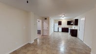 This single-floor space features an open-concept layout that seamlessly connects the living area to the kitchen, creating a functional and spacious feel. The flooring throughout is composed of light-toned square tiles, offering a clean and durable surface that ties the space together cohesively.

The kitchen is positioned at the far end of the open area and is equipped with dark espresso-toned cabinetry that provides a notable contrast against the light-colored walls. White appliances, including a refrigerator, stove, and under-counter sink setup, are included. The kitchen layout allows for efficient use of space while remaining visually accessible from the main living area.

The walls are finished in a soft, neutral beige tone, contributing to a bright and understated ambiance. Flush-mount ceiling light fixtures provide consistent overhead illumination throughout the space. An arched doorway leads to an adjacent room, suggesting additional living or sleeping quarters beyond the main open area. A secondary door, likely a utility or exterior access point, is also visible within the space.

Overall, the layout prioritizes open flow and practicality, making it a straightforward and versatile living environment.