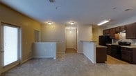 This single-floor residence features 3 bedrooms and 2 bathrooms within a well-organized layout designed for practical everyday living. The main living area is finished with neutral wall-to-wall carpeting and painted in warm, muted tones, creating a welcoming atmosphere throughout. Ceiling-mounted light fixtures provide consistent ambient lighting across the space.

The open-concept design connects the living area directly to the kitchen, which is defined by a raised breakfast bar counter with dark cabinetry and includes a full-size refrigerator. A second partial wall or bar structure on the opposite side of the living space adds visual interest and defines zones within the open floor plan.

A central hallway leads from the main living area toward the entry door, which is illuminated by a pendant light fixture, providing a clear and accessible path through the residence. The entry area transitions to tile or hard flooring, distinguishing it from the carpeted living zones.

Overall, the layout is efficient and straightforward, offering distinct functional areas within a cohesive single-level plan, suited for those seeking a manageable, low-maintenance living environment.