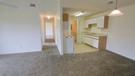 This single-floor residential unit offers a straightforward and functional layout designed for comfortable everyday living. The kitchen is positioned at the heart of the space, featuring white cabinetry with wood-tone accents, a dual-basin sink, a dishwasher, and a freestanding electric range and refrigerator. Light tile flooring in the kitchen area provides a clean, easy-to-maintain surface, while the countertops offer ample workspace for meal preparation. Overhead fluorescent lighting ensures the kitchen is well-lit and practical. Adjacent to the kitchen, the living area is finished with wall-to-wall carpet that extends down a central hallway, lending a warm and cohesive feel throughout the unit. The hallway connects to multiple rooms, each accessible via individual doors, suggesting a well-organized floor plan with clear separation between living and private spaces. The walls throughout are finished in a neutral, light tone, contributing to a bright and open atmosphere. Ceiling-mounted light fixtures provide consistent illumination across all areas. Overall, this unit presents a clean, move-in-ready space with a classic residential layout suited for a variety of occupants.