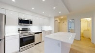 This thoughtfully designed one-bedroom space offers a clean, modern aesthetic across a single floor. The open-concept kitchen and living area serves as the heart of the space, showcasing sleek, high-gloss white cabinetry that extends to the ceiling for maximum storage. A striking white quartz-topped island anchors the kitchen, providing additional workspace and casual seating potential. The kitchen is fully equipped with a complete suite of stainless steel appliances, including a refrigerator, range, over-the-range microwave, and dishwasher, all complemented by a crisp white backsplash and recessed lighting overhead. Light-toned hardwood flooring flows seamlessly throughout, enhancing the bright, airy atmosphere. The living area is open and versatile, featuring a large window that allows natural light to fill the room while offering views of the surrounding urban streetscape. The bedroom is a well-proportioned private retreat, finished with the same light flooring and neutral white walls. It includes a built-in closet with mirrored sliding doors, adding both functionality and a sense of depth to the room. The overall design emphasizes clean lines, a neutral color palette, and modern finishes, creating a cohesive and contemporary living environment.