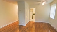 This single-floor residence offers a clean, functional layout with a warm and inviting ambiance throughout. The main living area is highlighted by rich hardwood floors that extend seamlessly across the space, complemented by soft neutral-toned walls that create a bright and airy feel. A ceiling fan with a light fixture is centrally positioned, providing both ventilation and ambient lighting. The open layout allows for flexible furniture arrangements, while white trim baseboards and interior doors add a crisp, classic finish to the space. An interior door with a round knob leads to an adjacent area, while a wide doorway opens directly into the kitchen. The kitchen features white cabinetry, a white refrigerator, and a patterned tile floor that contrasts neatly with the hardwood in the main room. The countertops include a dark-toned work surface, and the kitchen's bright, well-lit design makes it both practical and welcoming. The consistent use of white finishes and neutral walls throughout the space creates a cohesive and tidy aesthetic, making this a comfortable and move-in-ready single-story home.
