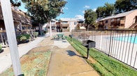 This residential community is composed of multiple multi-story stucco-exterior buildings arranged around a shared central courtyard, creating a cohesive and connected living environment. The buildings feature a warm tan and brown color palette with wood-accented balconies and railings, lending a classic, approachable architectural character to the community.

The outdoor common areas are a notable highlight, centered around a fenced swimming pool that serves as a social focal point. Adjacent to the pool is a dedicated BBQ and picnic area, complete with outdoor grills and round communal tables, making it well-suited for outdoor gatherings. Concrete walkways wind throughout the grounds, connecting the various buildings and amenity areas.

Mature trees — including large magnolias and shade trees — are distributed throughout the property, providing canopy coverage and a sense of established greenery. Well-maintained lawns and low shrubs line the building perimeters and pathways, contributing to the community's manicured appearance.

Each building features ground-floor and upper-floor units with sliding glass doors or windows opening to private balconies. Landscaping borders the building exteriors alongside HVAC units. The open sky and abundant natural light give the outdoor spaces a bright, airy feel throughout the day, making the communal areas inviting and functional for residents year-round.