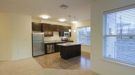 This single-floor residence showcases an open-concept layout that seamlessly connects the kitchen and living areas. The kitchen is a standout feature, appointed with rich, dark espresso cabinetry that extends to the ceiling for ample storage. Stainless steel appliances, including a refrigerator, range, microwave, and dishwasher, provide a sleek, modern aesthetic. A central island with a dark finish offers additional counter space and casual seating potential. Pendant lighting hangs above the island, complementing the flush-mount ceiling fixtures that illuminate the space evenly. Light granite-toned countertops contrast beautifully against the dark cabinetry. Hardwood-style flooring runs throughout the kitchen and transitions into carpeting in the adjacent living area. Large windows along the kitchen wall allow substantial natural light to fill the space, offering views of the surrounding exterior. The neutral, warm wall tones and clean lines throughout contribute to a contemporary yet inviting ambiance well-suited for both everyday living and entertaining.