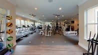This spacious fitness center is designed to accommodate a full range of workout needs within a single-floor layout. The space is organized into distinct functional zones, beginning with an entry area finished with dark carpeting, crown molding, and French doors with glass panes that provide a welcoming transition into the facility. Emergency safety signage and posted fitness center policies are clearly displayed near the entrance.

The main workout area is generously proportioned, featuring dark carpeting throughout the strength training zones and hard flooring in the cardio and stretching areas. Multiple ceiling fans are evenly distributed across the ceiling alongside recessed lighting, ensuring consistent air circulation and illumination throughout. Large windows line the perimeter walls, flooding the space with natural light and offering views of the surrounding greenery.

The strength training section is well-stocked with multiple cable machines, weight benches, and a variety of resistance equipment, complemented by wall-mounted mirrors that enhance the sense of space. The cardio zone includes treadmills, elliptical trainers, and stationary bikes, all positioned to take advantage of the outdoor views. A dedicated area houses accessories including medicine balls, resistance bands, and yoga mats on a rack. Wall-mounted televisions provide entertainment throughout.