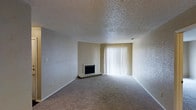 This single-floor space includes a living room that serves as its central feature. The room is finished with wall-to-wall carpeting in a neutral tone, contributing to a warm and understated aesthetic. A fireplace with a raised hearth sits along one wall, offering a functional focal point for the space. Adjacent to the fireplace, a large sliding glass door fitted with vertical blinds allows abundant natural light to fill the room, creating a bright and open atmosphere. The walls are finished in a light, neutral palette, complementing the overall subdued color scheme. A textured popcorn ceiling runs throughout the room, a characteristic finish commonly found in residential spaces of this style. The layout is open and unobstructed, providing flexibility for furniture arrangement. Neutral-toned walls and baseboards frame the space cleanly, while the combination of the fireplace and the large glass door makes the living area both functional and inviting. The single-floor layout ensures convenient accessibility throughout the space.