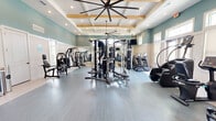 Located at 12525 Tibbets Lane in Jacksonville, Florida, this fitness center offers a generously sized, single-floor layout designed to support a full range of workout activities. The space makes a strong architectural impression with its soaring coffered ceilings accented by warm wood beams and dramatic oversized ceiling fans that enhance both airflow and visual appeal. Recessed lighting throughout ensures the space is bright and welcoming at all hours.

The interior is finished in a calming palette of soft teal walls, white wainscoting, and light-toned resilient flooring, creating a clean and motivating environment. Large multi-pane windows along the perimeter flood the space with natural light and offer pleasant views of the surrounding landscaped grounds, including palm trees visible from the main workout area.

The facility is well-appointed with a wide variety of equipment, including a multi-station cable machine, selectorized strength machines, a dumbbell rack, FitBench workout stations, treadmills, stair climbers, ellipticals, a stationary bike, and a rowing machine. Wall-to-wall mirrors in key zones enhance spatial depth and support form-checking during workouts. A mounted flat-screen television and direct exterior access via glass-paned doors round out this polished and fully functional fitness facility.