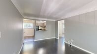 This single-floor unit offers three bedrooms and one bathroom within a well-organized layout. The living room is a standout space, featuring dark-stained hardwood floors, soft gray walls, and large windows fitted with vertical blinds that allow generous natural light to fill the room. Crown molding adds a subtle architectural detail to the ceiling.

The kitchen connects to the living area via a pass-through design with a granite-style countertop bar, creating an open sightline between the two spaces. The kitchen is equipped with gray cabinetry, a gas range, stainless sink, and a full-size refrigerator, with tile flooring and a window overlooking greenery outside.

The bedrooms are carpeted and neutral in tone, each with floor vents and closet access. One closet features wire shelving for added storage utility. The primary bedroom benefits from a large window with views of outdoor greenery.

The bathroom is cleanly finished with dark tile flooring, a wood-toned vanity with a white countertop, and a full tub-shower combination surrounded by white subway tile. A small window above the tub provides natural ventilation. A hallway connects all rooms, contributing to a straightforward and practical floor plan.