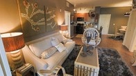 This single-floor space showcases a thoughtfully designed open-concept layout that seamlessly blends the living, dining, and kitchen areas into one cohesive environment. The living area is anchored by a plush, neutral-toned sofa accented with patterned throw pillows, set atop a rich, dark shag area rug that adds warmth and texture to the hardwood-style flooring beneath. A striped storage trunk doubles as a coffee table, contributing both function and charm. Decorative wall art in a triptych format adds a bold artistic focal point, while warm ambient lighting from table lamps and track lighting creates an inviting, intimate atmosphere. A distinctive white rattan chair introduces a touch of eclectic style. The dining area features a round table with elegant seating, positioned near the kitchen for easy flow. The kitchen is equipped with dark appliances, including a black stove and microwave, complemented by warm wood cabinetry and ample counter space. The earthy, neutral color palette — featuring warm taupes, grays, and browns — runs throughout the space, tying together the various design elements into a unified, welcoming aesthetic. The open layout enhances the overall sense of space and accessibility.