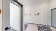 Located at 1818 N 53rd St in Philadelphia, PA, this single-floor commercial space offers a thoughtfully designed layout comprising two hallways and a private office. The main hallway is a standout feature, characterized by soaring ceilings, light wood flooring, and a generous wall of floor-to-ceiling windows that flood the space with abundant natural light and offer views of the surrounding neighborhood streetscape. A row of wooden desks with modern chairs lines the windowed wall, complemented by floating shelves displaying greenery and décor. Richly patterned area rugs add warmth and texture throughout. A secondary hallway connects to a staircase with a carpeted tread and metal handrails, alongside restroom access and an exit, ensuring practical circulation throughout the space. The private office is an intimate, focused room furnished with a white conference table, green upholstered chairs, and a wall-mounted flat-screen display, making it ideal for meetings or focused work sessions. Clean white walls, recessed lighting, and consistent blue-gray baseboard trim tie the space together with a cohesive, modern aesthetic. The overall ambiance is bright, inviting, and professionally appointed.