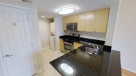 This single-floor space showcases a well-appointed kitchen as its centerpiece, designed with both functionality and style in mind. The kitchen features striking dark granite countertops that provide ample prep space, complemented by light maple wood cabinetry that extends to the upper walls for generous storage capacity. Stainless steel appliances, including a gas range with oven, built-in microwave, and a undermount stainless steel sink, lend a sleek, contemporary aesthetic to the space.

The kitchen island or peninsula with matching dark granite offers additional workspace and a natural gathering point. Light-toned tile flooring runs throughout, creating a clean, cohesive look that brightens the space. Recessed and flush-mount ceiling lighting ensures the kitchen is well-illuminated throughout the day.

A notable practical feature is the dedicated in-unit laundry area, visible just beyond the kitchen, housing a stacked washer and dryer unit within a discreet alcove. The open layout of the kitchen connects seamlessly to the adjacent hallway, contributing to an efficient and accessible floor plan throughout the home.